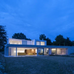 A modern two-story house with large glass windows and a flat roof, set in a landscaped yard under a twilight sky. The light from the interior creates a warm ambiance, and trees frame the background.