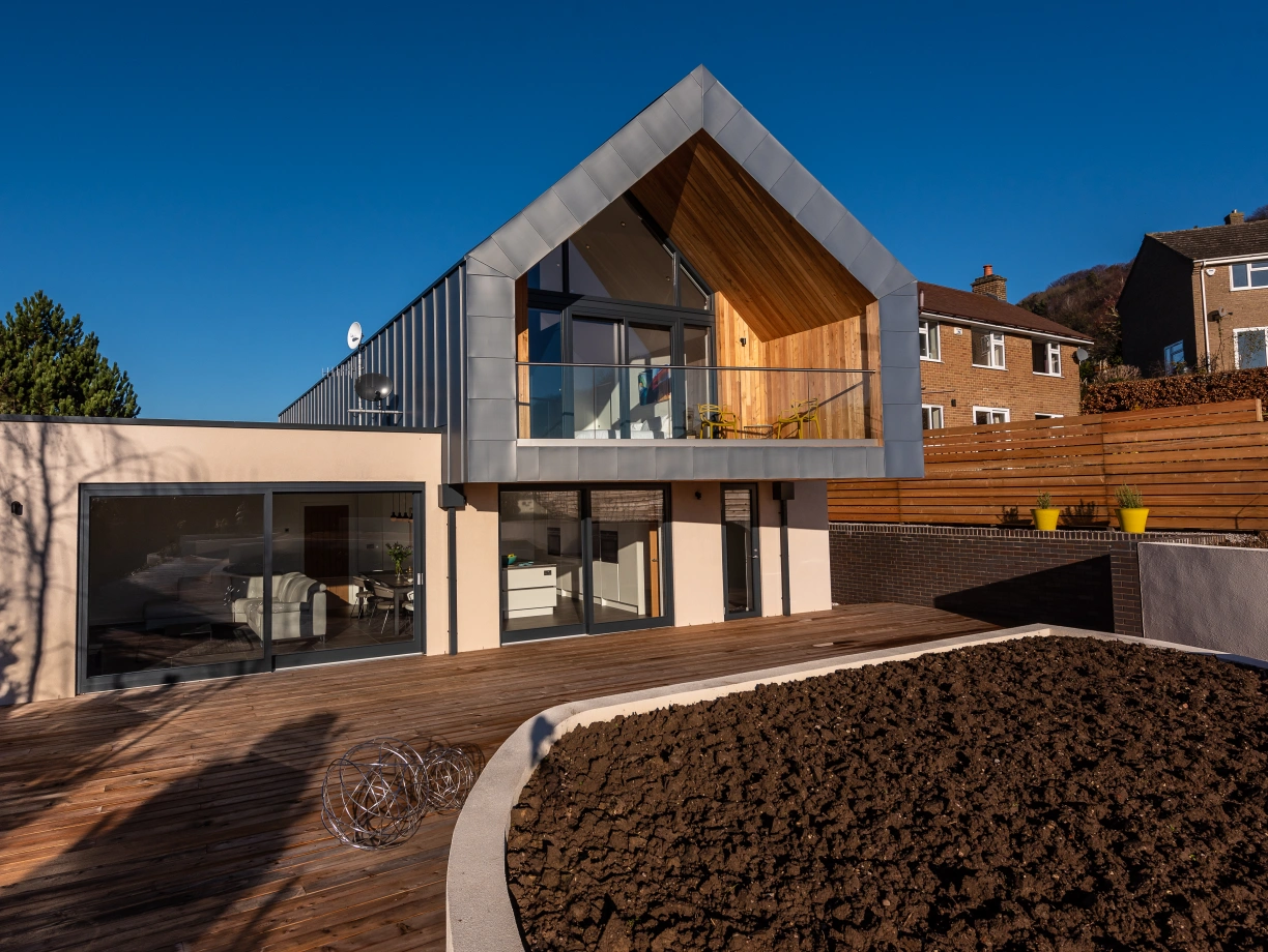 A modern two-story house features a large wooden deck, a sloping roofline, and expansive glass windows. The exterior combines light-colored walls with natural wood accents, set against a clear blue sky. A landscaped area with dark soil is visible in the foreground.