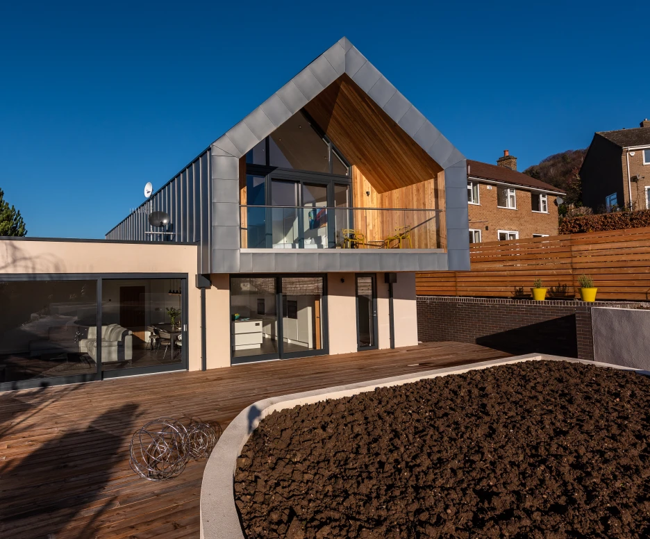 A modern two-story house features a large wooden deck, a sloping roofline, and expansive glass windows. The exterior combines light-colored walls with natural wood accents, set against a clear blue sky. A landscaped area with dark soil is visible in the foreground.