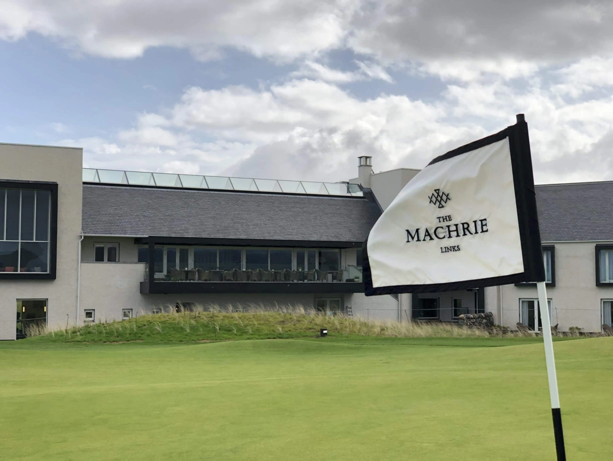 The Machrie Links flag stands in front of a modern golf clubhouse under a partly cloudy sky. The building features large windows and an outdoor seating area.