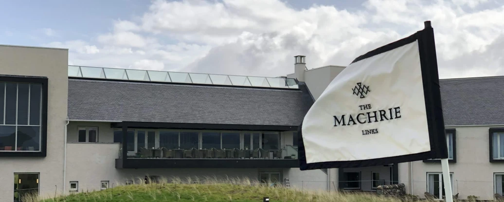 A green golf course with a black and white flag reading "The Machrie Links" in the foreground. In the background, a modern building with large windows and a terrace is visible under a cloudy sky.