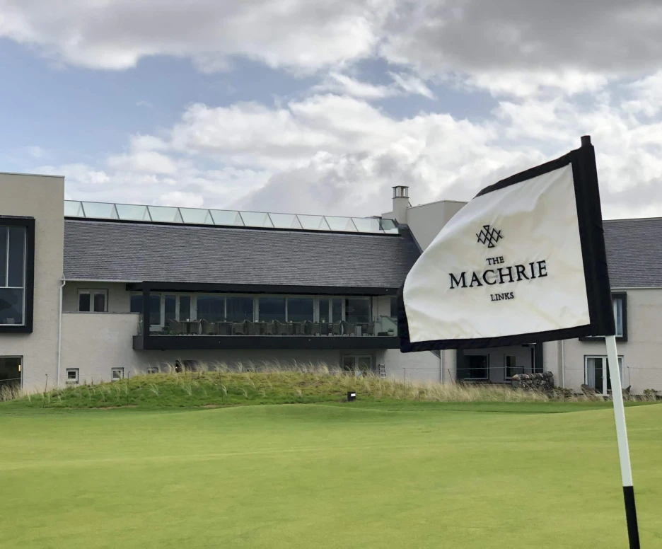 A golf flag with "The Machrie Links" logo stands in front of a modern clubhouse featuring large windows and a balcony, set against a cloudy sky.