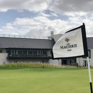 A green golf course with a black and white flag reading "The Machrie Links" in the foreground. In the background, a modern building with large windows and a terrace is visible under a cloudy sky.