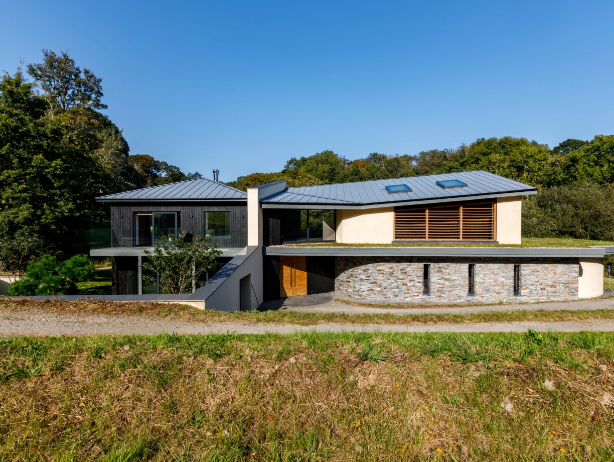 Modern architectural house featuring a mix of wood and stone materials, large windows, and a unique curved roof design. The home is set against a backdrop of lush greenery under a clear blue sky. A gravel path leads up to the entrance.