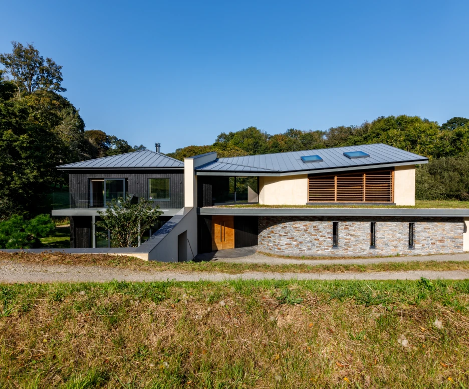 Modern architectural house featuring a mix of wood and stone materials, large windows, and a unique curved roof design. The home is set against a backdrop of lush greenery under a clear blue sky. A gravel path leads up to the entrance.
