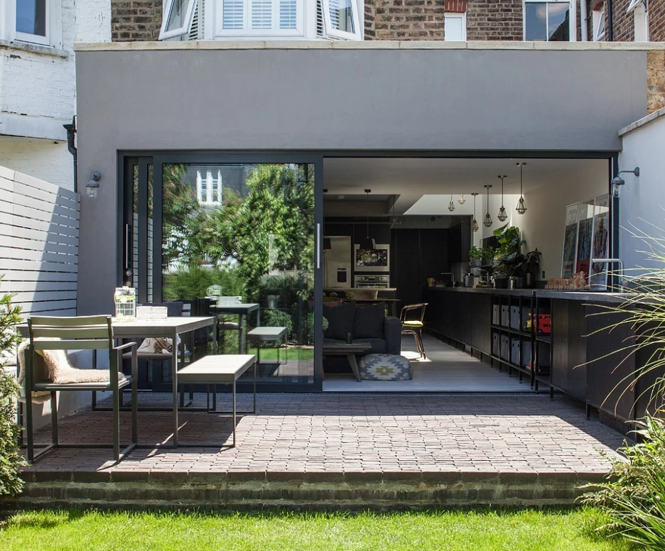 A modern outdoor patio features a sleek dining table with chairs, surrounded by lush greenery. A large glass door opens to a contemporary interior space, showcasing a minimalist design with dark cabinetry and bright decor. Sunlight illuminates the area, enhancing the inviting atmosphere.