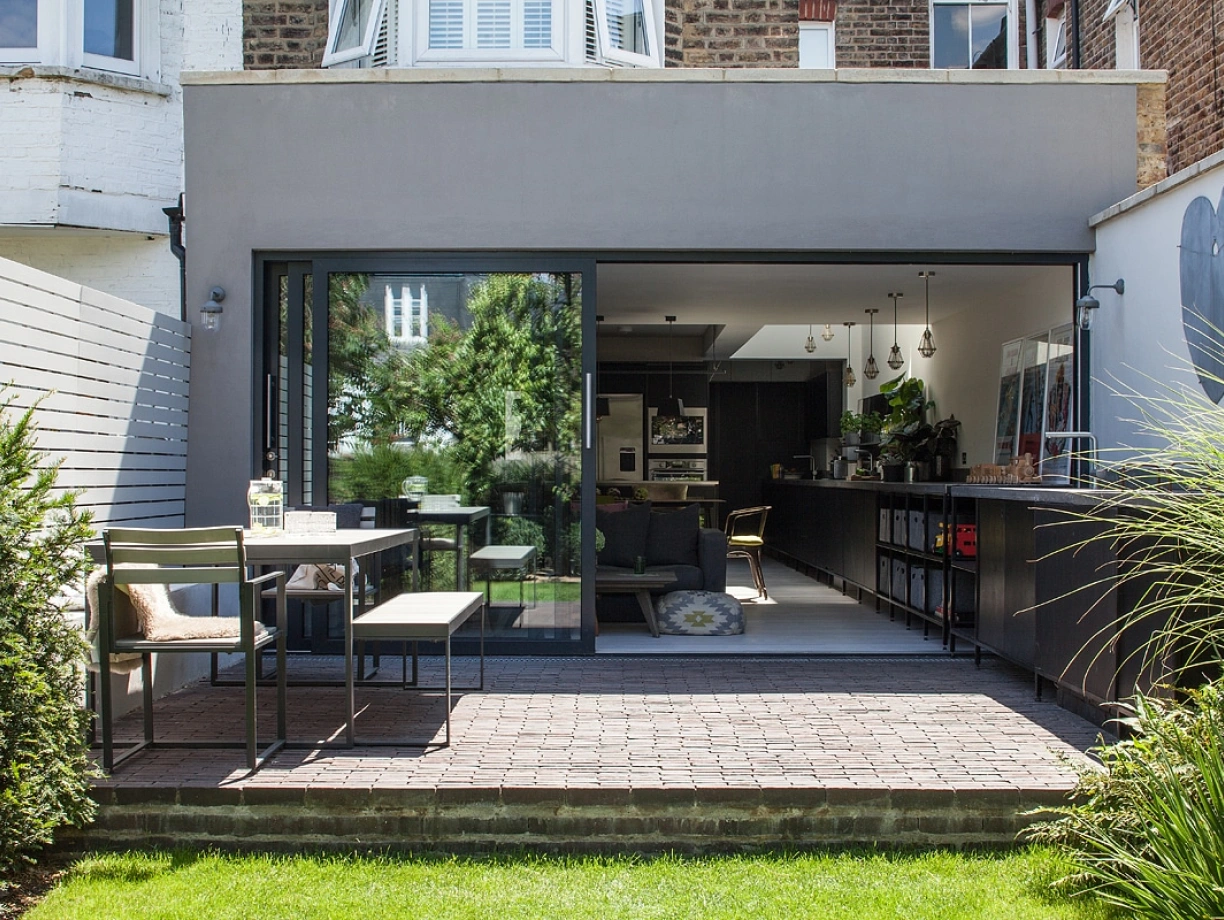 A modern outdoor patio features a sleek dining table with chairs, surrounded by lush greenery. A large glass door opens to a contemporary interior space, showcasing a minimalist design with dark cabinetry and bright decor. Sunlight illuminates the area, enhancing the inviting atmosphere.