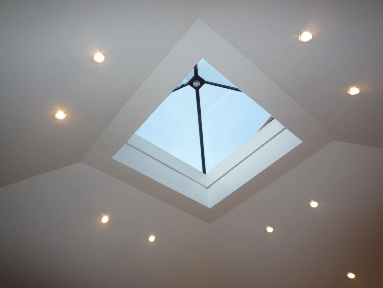 Plafond lumineux avec un puits de lumière au centre, entouré de lumières encastrées. Ciel bleu visible à travers la fenêtre en verre.