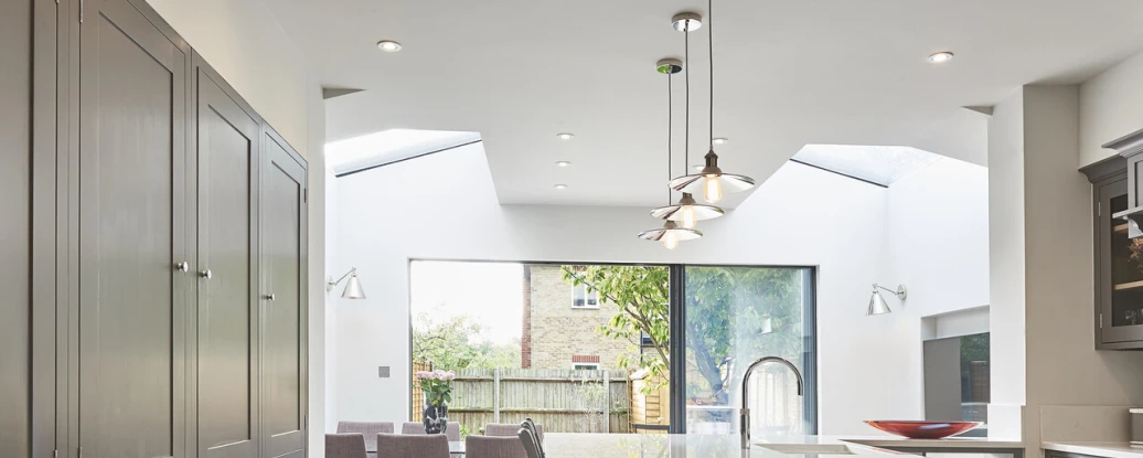 A modern kitchen featuring gray cabinetry, an island with a countertop, and large windows that allow natural light. A dining area with purple chairs is visible in the background, alongside a view of greenery outside.