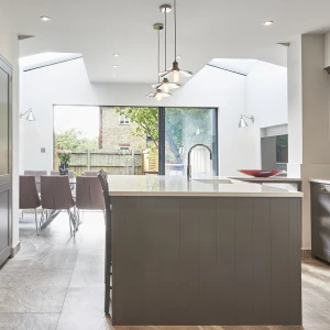 A modern kitchen featuring gray cabinetry, an island with a countertop, and large windows that allow natural light. A dining area with purple chairs is visible in the background, alongside a view of greenery outside.