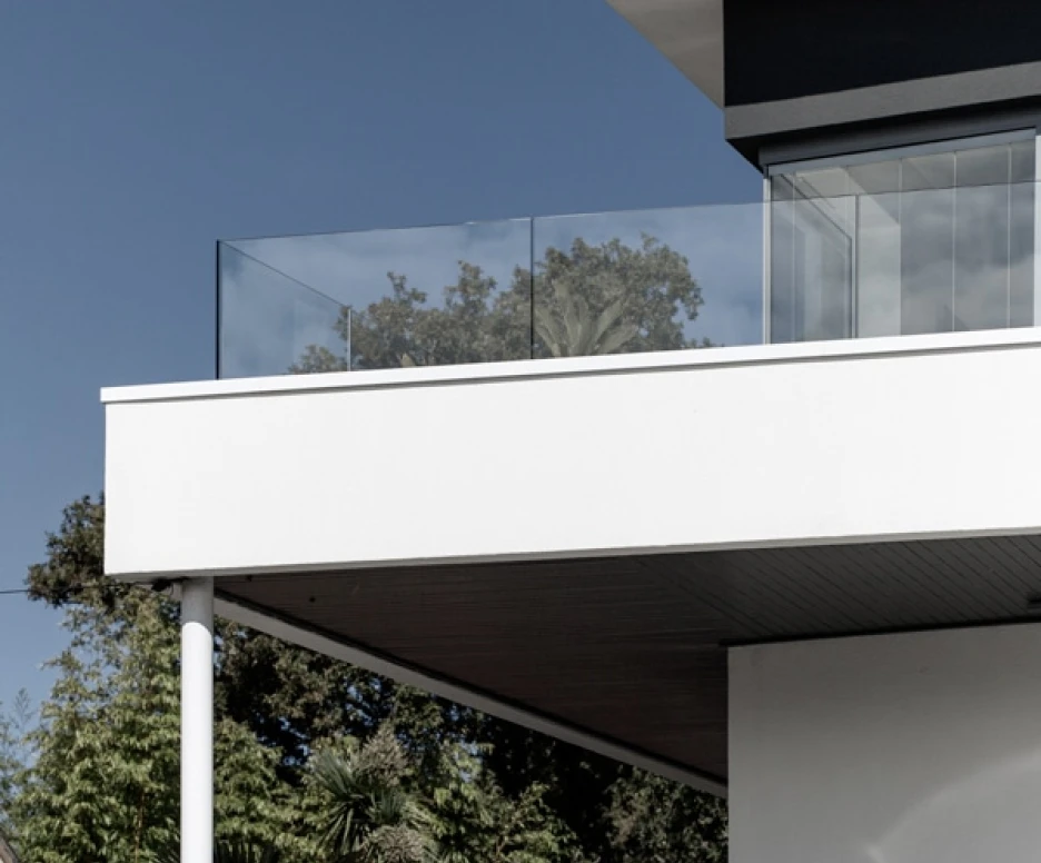 Modern architecture featuring a stylish upper balcony with glass railing, set against a clear blue sky. Green trees and plants surround the base of the building.