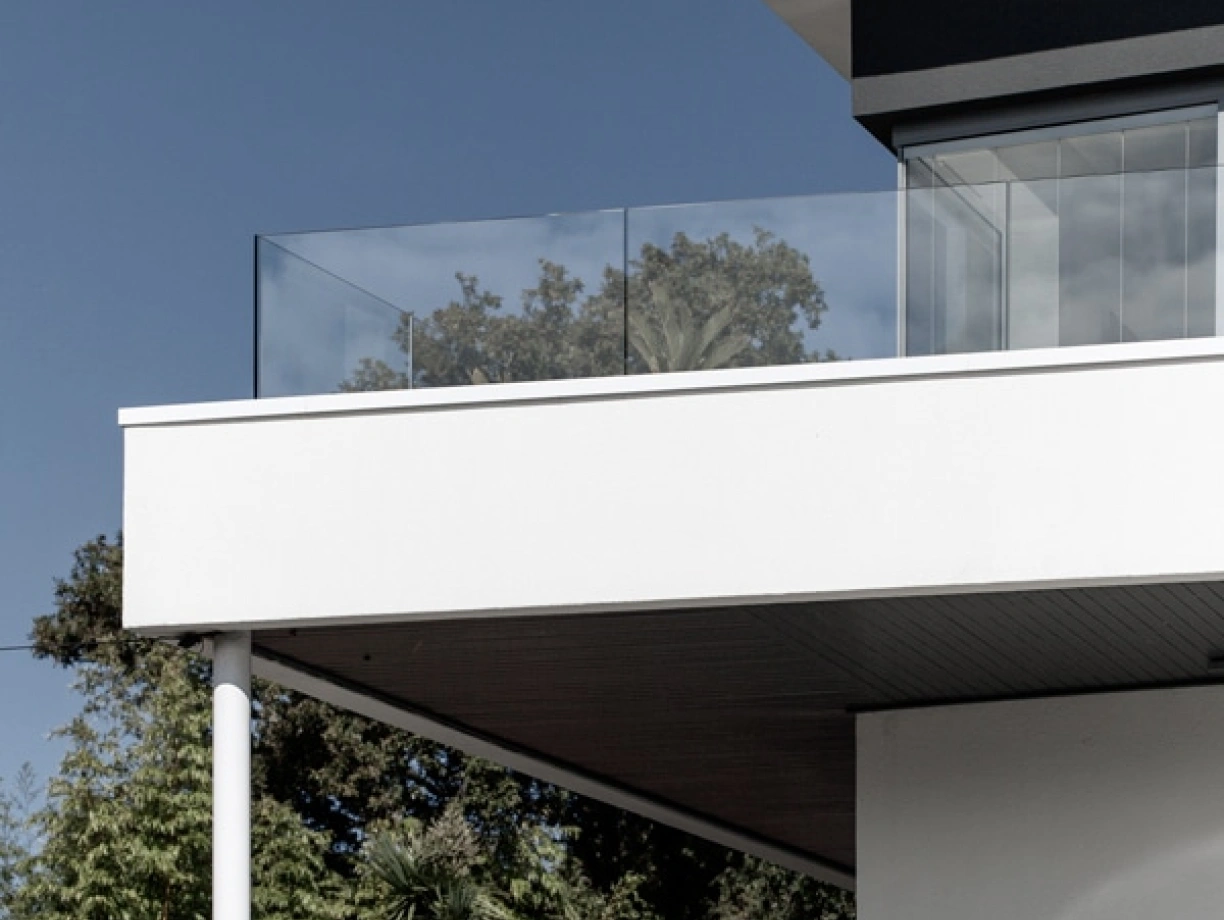 Modern architecture featuring a stylish upper balcony with glass railing, set against a clear blue sky. Green trees and plants surround the base of the building.