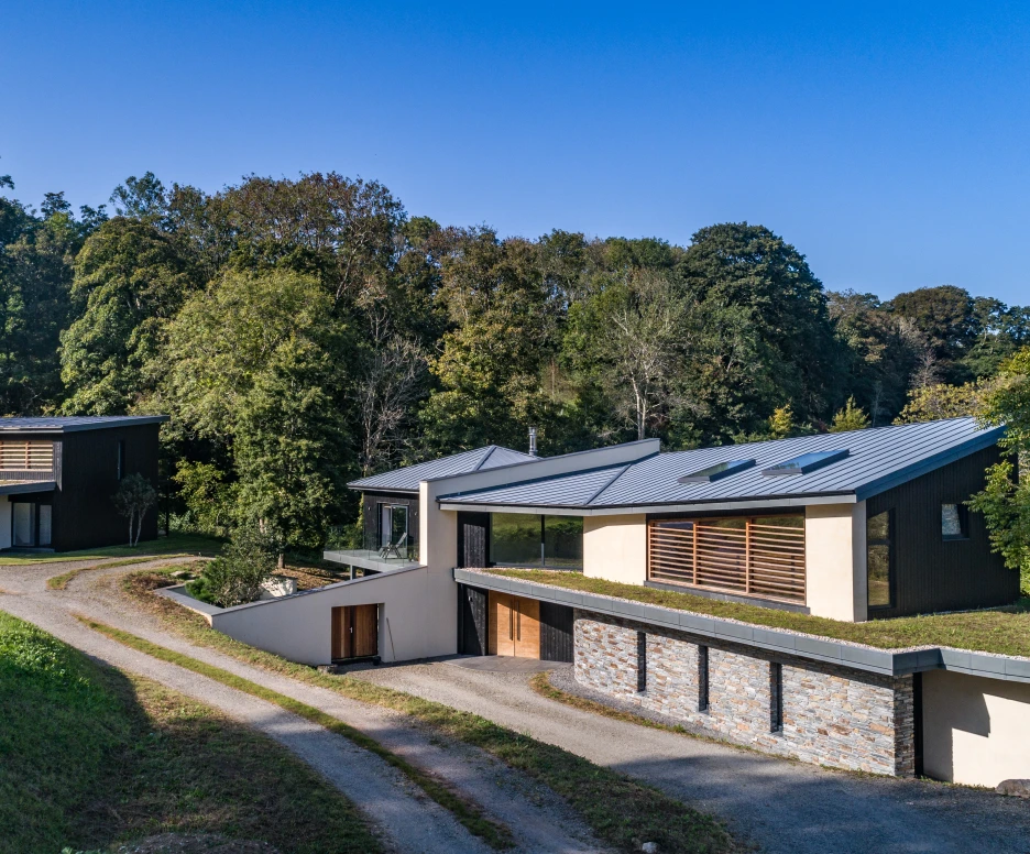 Two modern houses nestled in a green landscape with trees. One house features a sloped roof and large windows, while the other has a flat roof and stone accents. A gravel path leads up to the homes. Clear blue sky overhead.