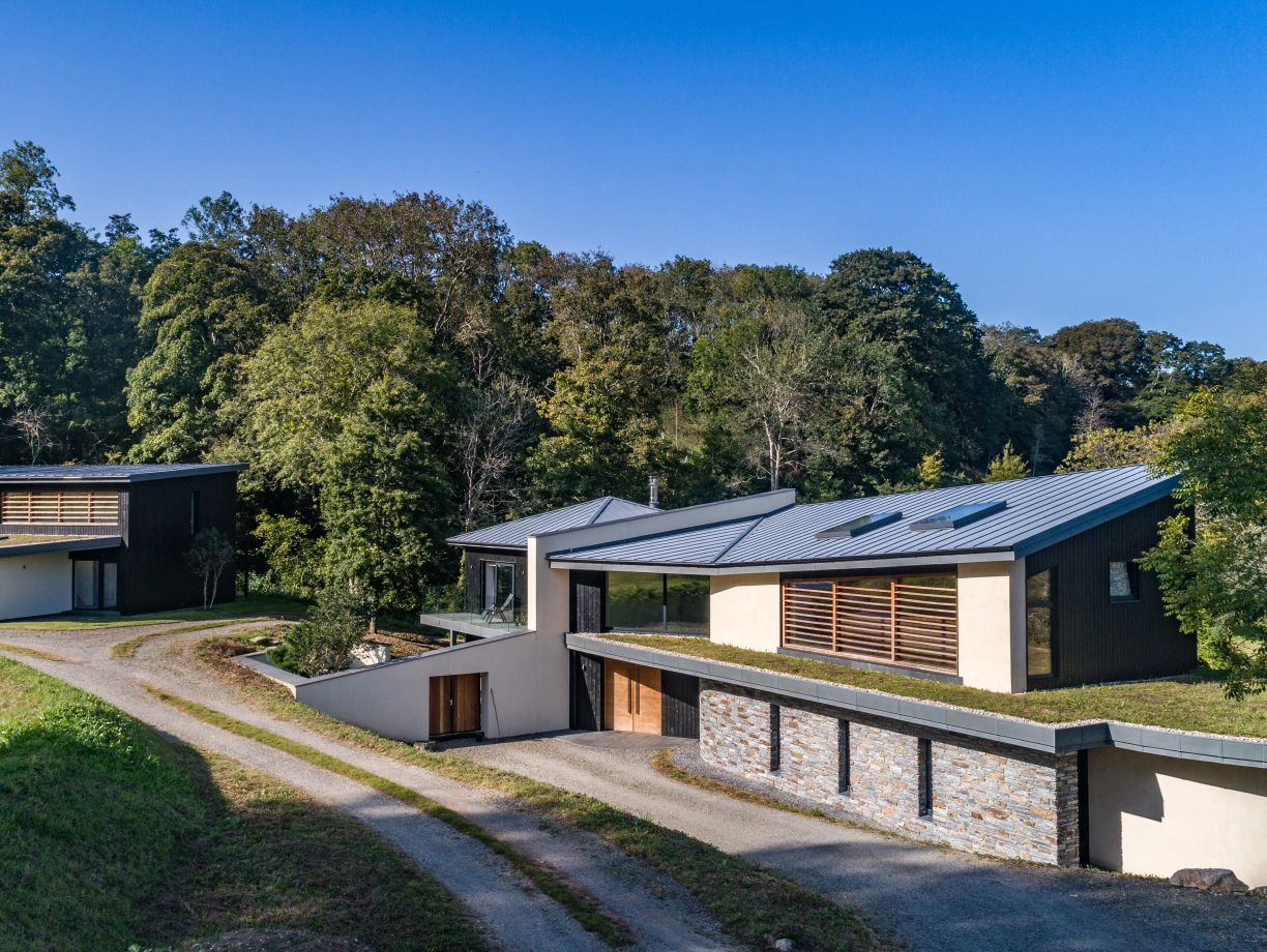 Two modern houses nestled in a green landscape with trees. One house features a sloped roof and large windows, while the other has a flat roof and stone accents. A gravel path leads up to the homes. Clear blue sky overhead.
