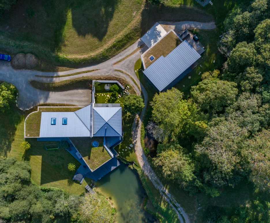 Aerial view of two modern buildings surrounded by lush greenery and a small pond. One building features a green roof with skylights, while the other has a sleek design. Winding paths connect them, with a clear blue sky above.