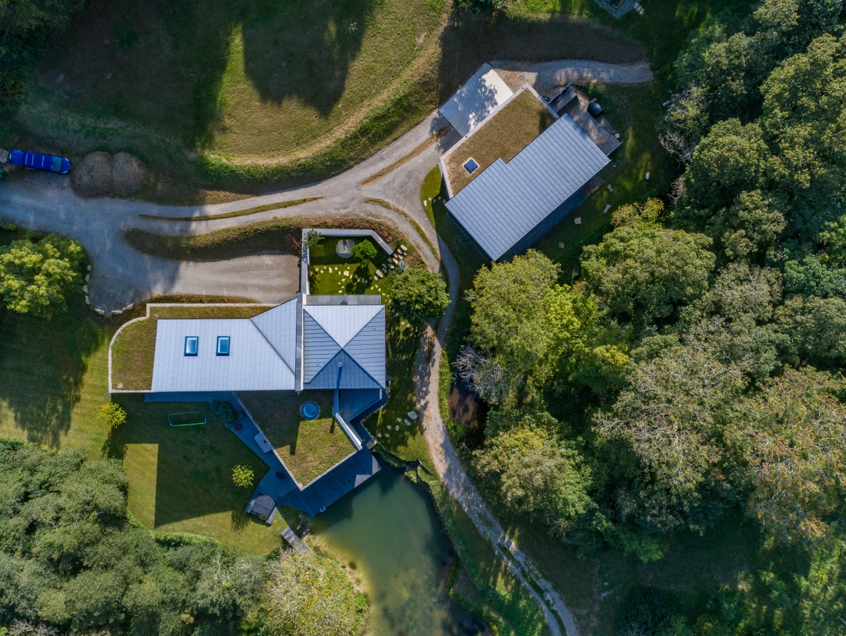 Aerial view of two modern buildings surrounded by lush greenery and a small pond. One building features a green roof with skylights, while the other has a sleek design. Winding paths connect them, with a clear blue sky above.