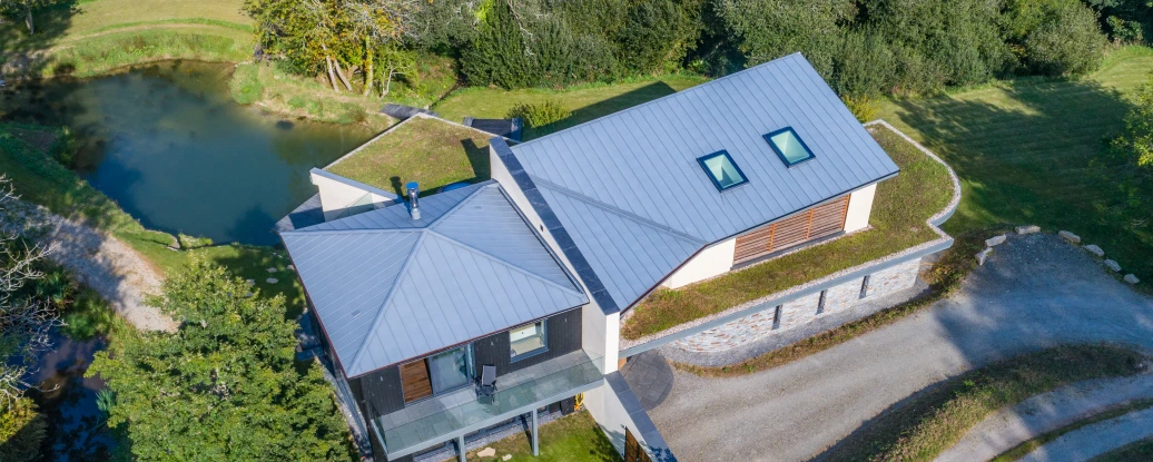Aerial view of a modern two-story house with a gray metal roof, surrounded by trees and a small pond. A gravel driveway leads to the home, which features a green lawn and landscaped garden.