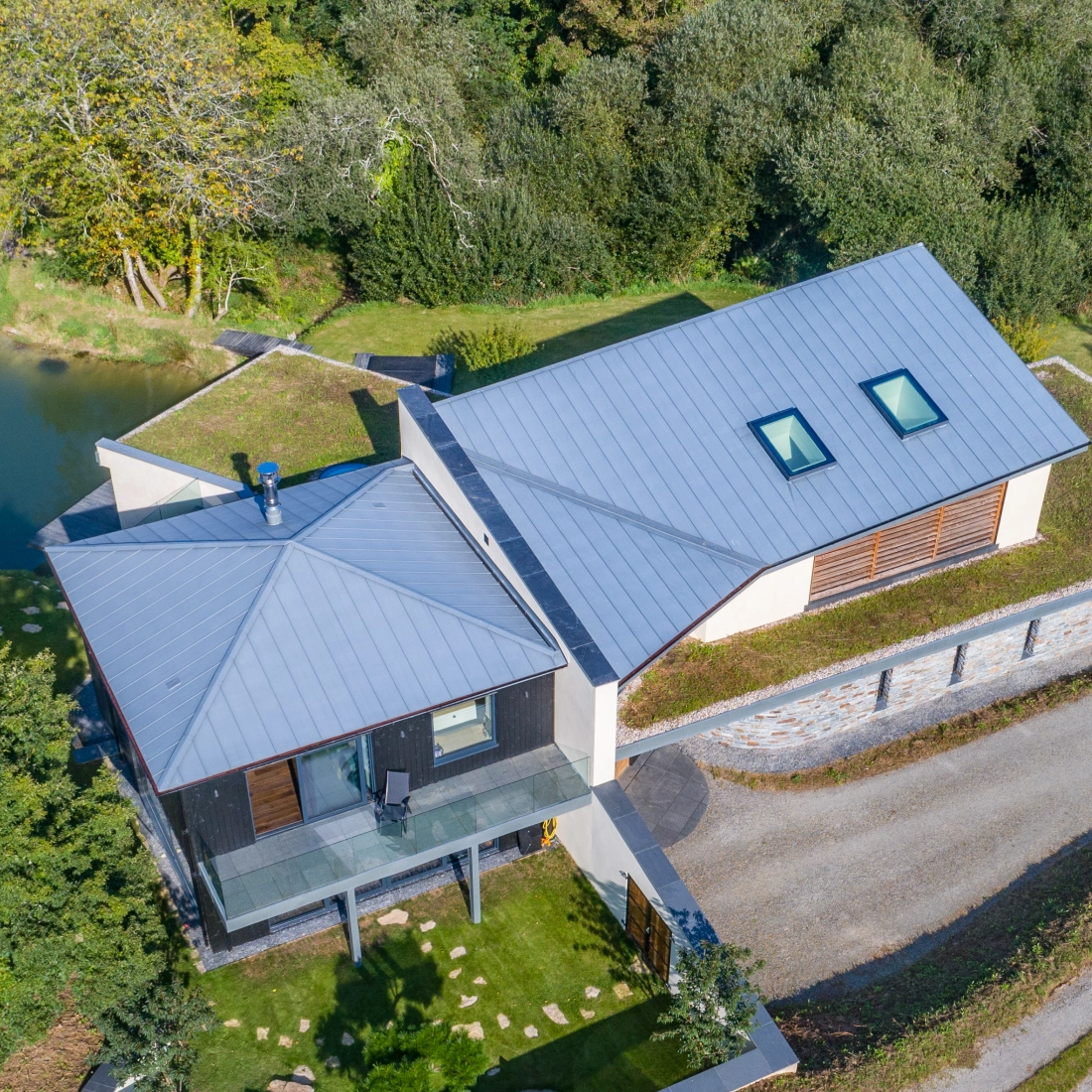 Aerial view of a modern two-story house with a gray metal roof, surrounded by trees and a small pond. A gravel driveway leads to the home, which features a green lawn and landscaped garden.