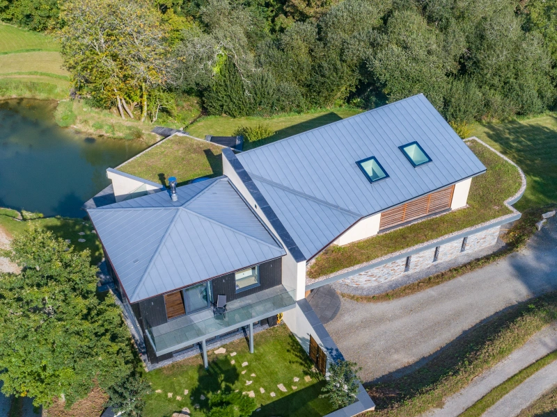 Aerial view of a modern two-story house with a gray metal roof, surrounded by trees and a small pond. A gravel driveway leads to the home, which features a green lawn and landscaped garden.