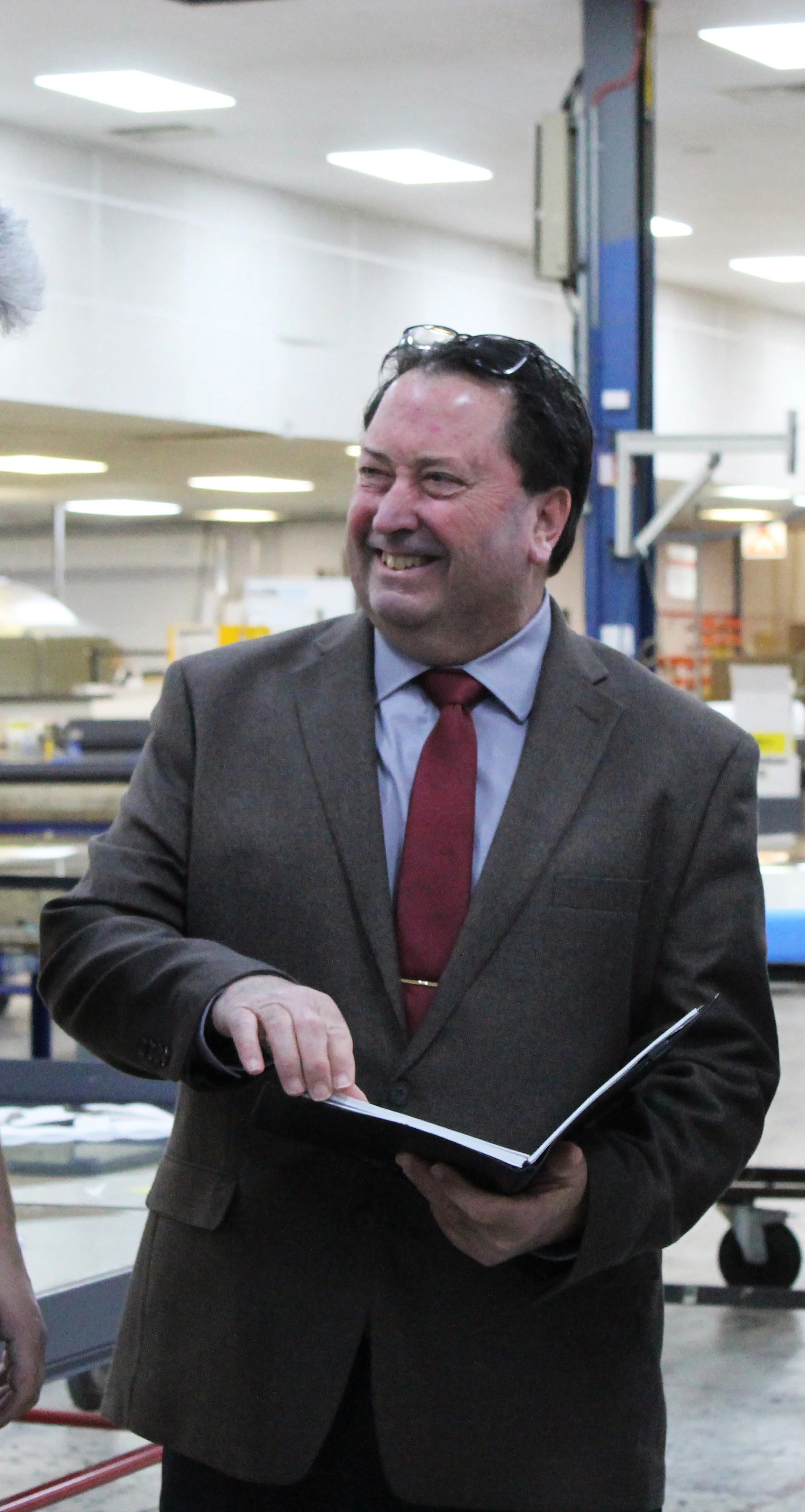 A man in a brown suit and red tie smiles while holding an open book in a factory setting. He appears engaged in conversation, surrounded by industrial equipment.