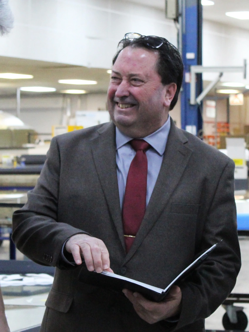 A man in a brown suit and red tie smiles while holding an open book in a factory setting. He appears engaged in conversation, surrounded by industrial equipment.