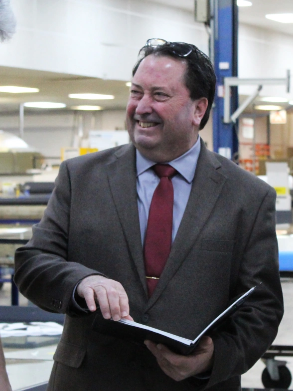 A man in a brown suit and red tie smiles while holding an open book in a factory setting. He appears engaged in conversation, surrounded by industrial equipment.