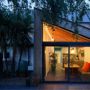 A modern home extension featuring large glass doors, wooden beams, and warm lighting. The interior includes a dining area with a wooden table, plants, and a cozy seating space. Surrounding the house are trees and a garden.