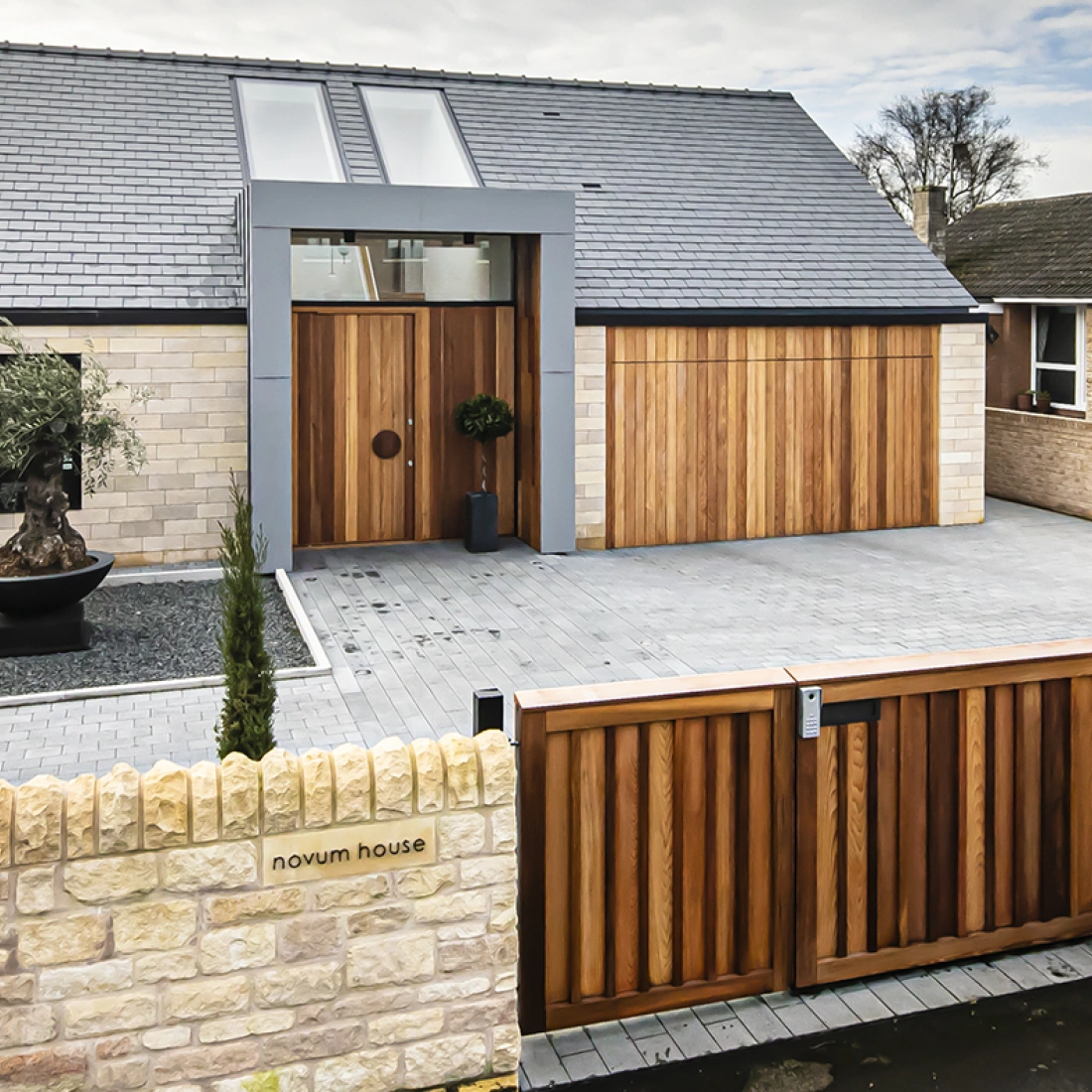 A modern house with a sleek design features a large wooden front door and a matching garage door. The exterior is a combination of stone and gray cladding, surrounded by a landscaped driveway with small trees and a decorative planter. A stone wall and wooden gate mark the entrance, labeled "Novum House."