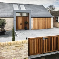 Casa moderna con fachada de piedra, tejado gris y puertas de madera. La entrada cuenta con un jardín minimalista, una planta decorativa y una fuente. Un portón de madera da acceso al patio.