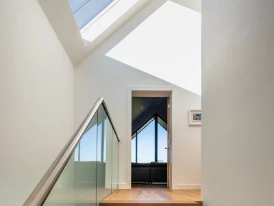 A modern staircase leading to a room with large, angular windows. Natural light floods the space from a skylight above, highlighting the wooden steps and glass railing. A doorway at the top opens to a bright living area with a view outside.