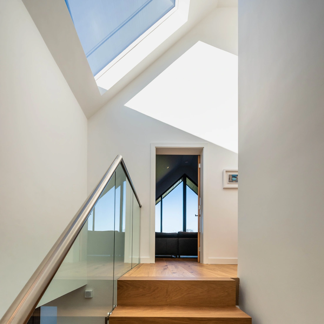 A modern staircase leading to a room with large, angular windows. Natural light floods the space from a skylight above, highlighting the wooden steps and glass railing. A doorway at the top opens to a bright living area with a view outside.