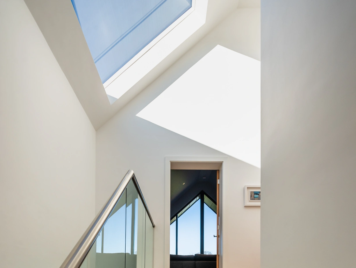 Wooden stairs lead upward into a well-lit hallway with a skylight. A contemporary glass railing complements the modern design. A doorway at the top reveals a room with large windows, offering a view of the outdoors.