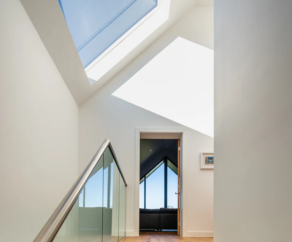 Wooden stairs lead upward into a well-lit hallway with a skylight. A contemporary glass railing complements the modern design. A doorway at the top reveals a room with large windows, offering a view of the outdoors.