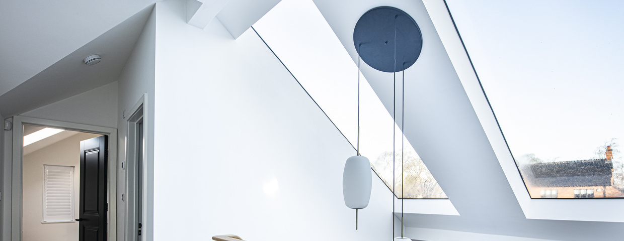 Bright, modern interior featuring a spacious hallway with a skylight and minimalist design. Light walls and a wooden railing complement the contemporary aesthetic. A circular pendant light hangs from the ceiling, illuminating the space.