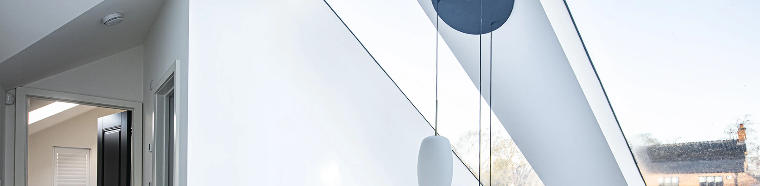 Bright, modern interior featuring a spacious hallway with a skylight and minimalist design. Light walls and a wooden railing complement the contemporary aesthetic. A circular pendant light hangs from the ceiling, illuminating the space.