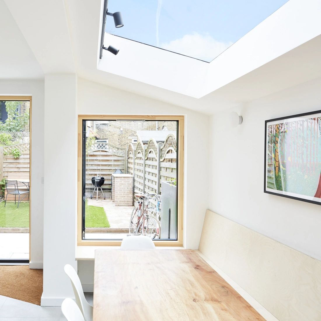 A modern kitchen-dining area features a light wood table and a vase of fresh flowers. Large windows provide views of a landscaped garden with a grassy area and patio. A skylight adds natural light to the space.