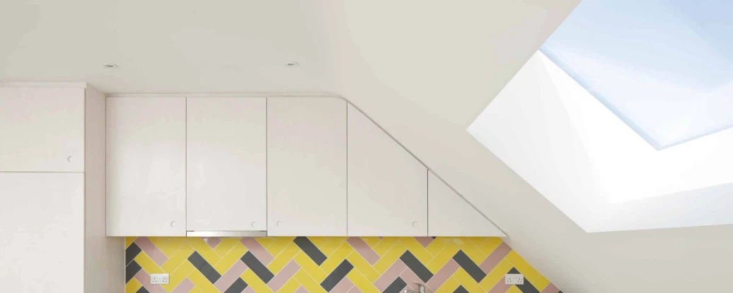 Modern kitchen featuring a sloped ceiling, white cabinetry, and a vibrant yellow, pink, and black herringbone tile backsplash. A stainless steel stove is positioned against the wall, with a skylight above allowing natural light to fill the space.