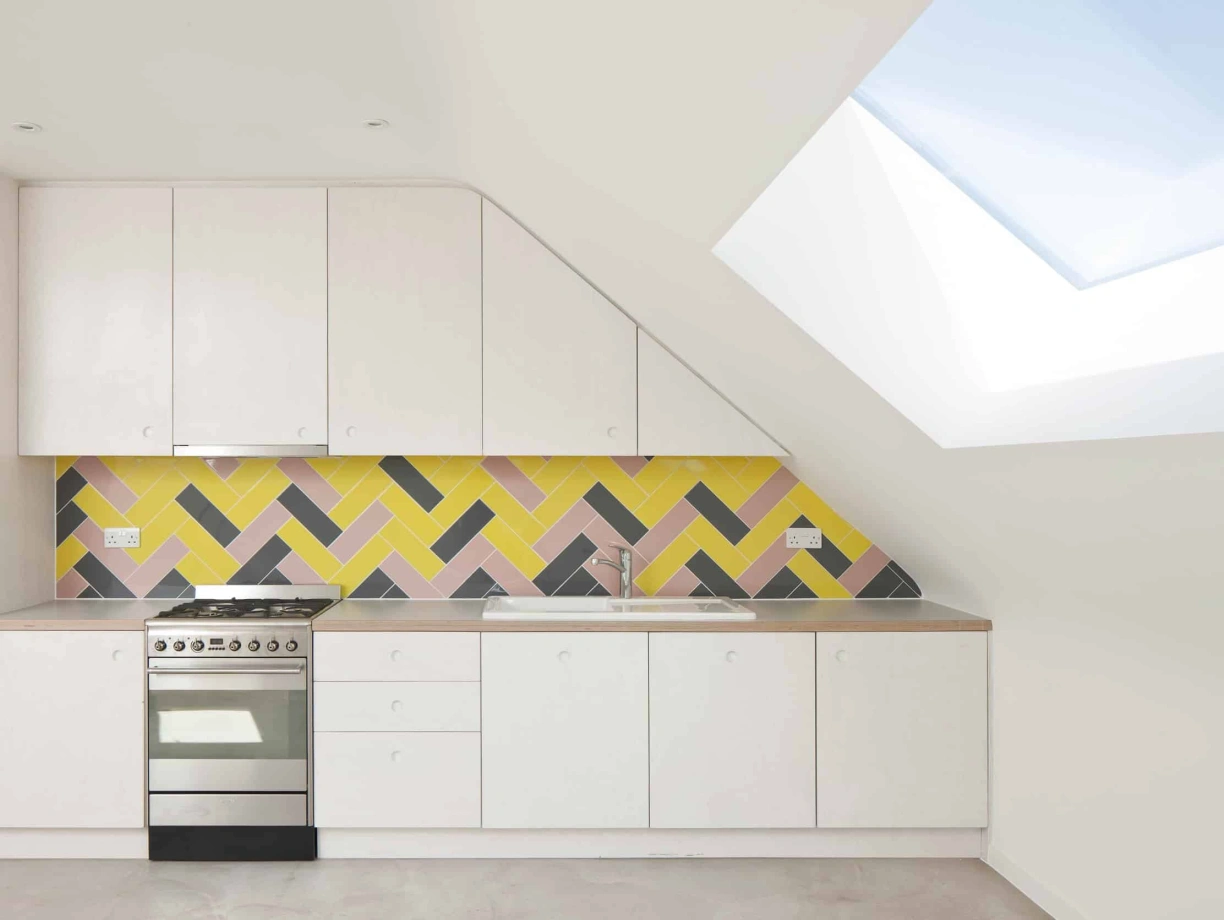 Modern kitchen featuring a sloped ceiling, white cabinetry, and a vibrant yellow, pink, and black herringbone tile backsplash. A stainless steel stove is positioned against the wall, with a skylight above allowing natural light to fill the space.