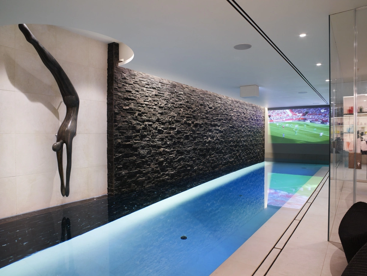 A sleek indoor swimming pool with a modern design. A stone wall features a sculpture of a diver, while a screen displays a sports game in the background. The pool has clear blue water and contemporary lighting.