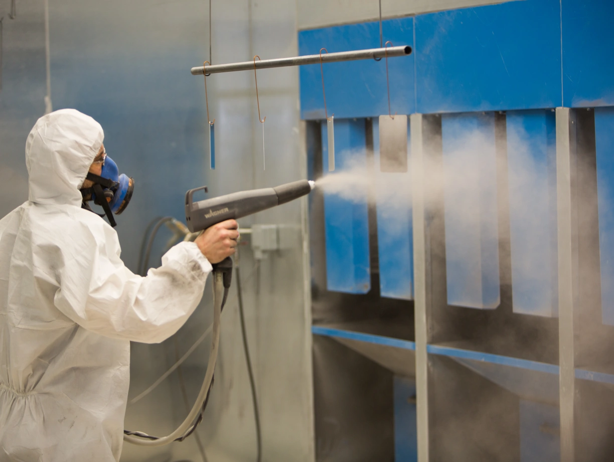 A person in a protective suit and mask uses a spray gun to apply a coating inside a spray booth. The background features blue panels and a mist of spray particles.