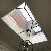 A modern interior featuring a skylight with a geometric black frame. A stylish black and glass chandelier hangs below the skylight, illuminating the space. Elegant white paneling surrounds the area.