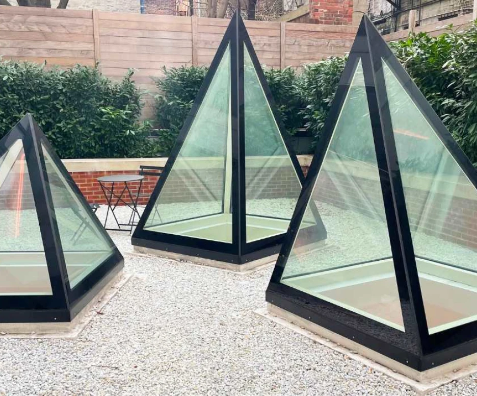Three glass pyramids with black frames are situated in a gravel area surrounded by lush greenery and a wooden fence. A small black table and chairs are visible nearby.
