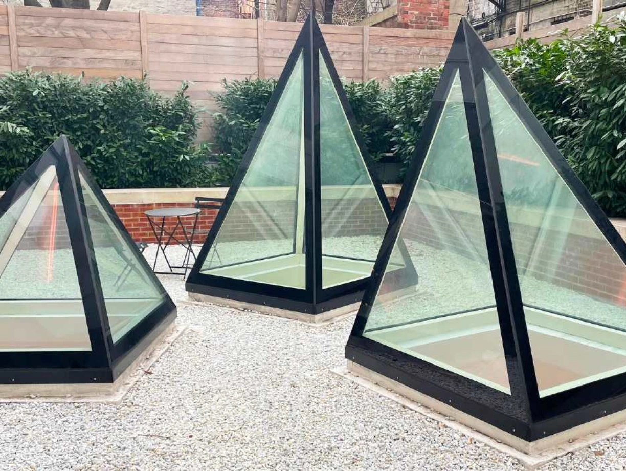 Three glass pyramids with black frames are situated in a gravel area surrounded by lush greenery and a wooden fence. A small black table and chairs are visible nearby.