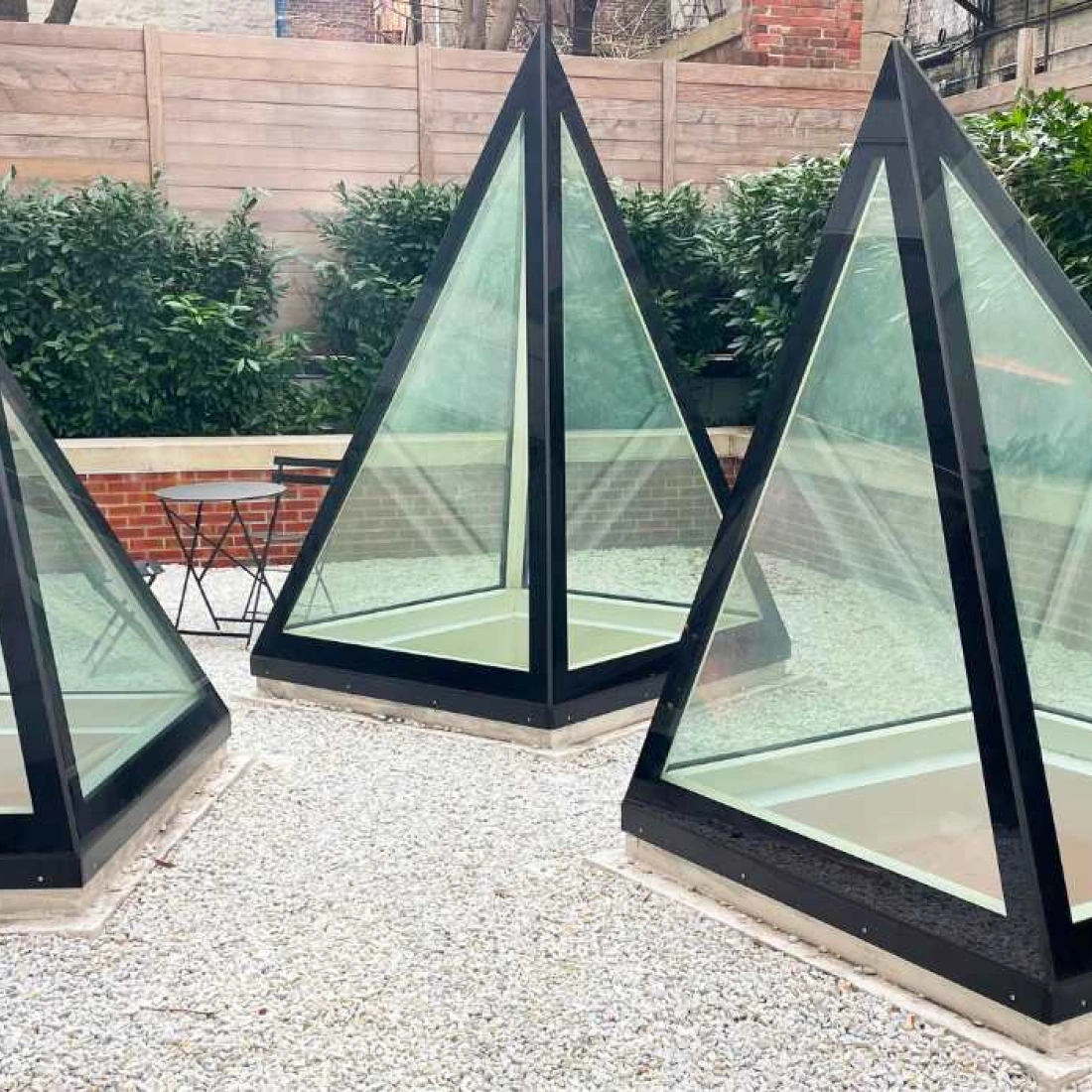 Trois pyramides en verre et métal noir, disposées sur un sol en gravier, entourées de verdure et de clôtures en bois.