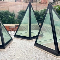 Three glass and black-framed pyramids set in a gravel area surrounded by greenery. The pyramids are arranged at different angles, creating a modern architectural design.