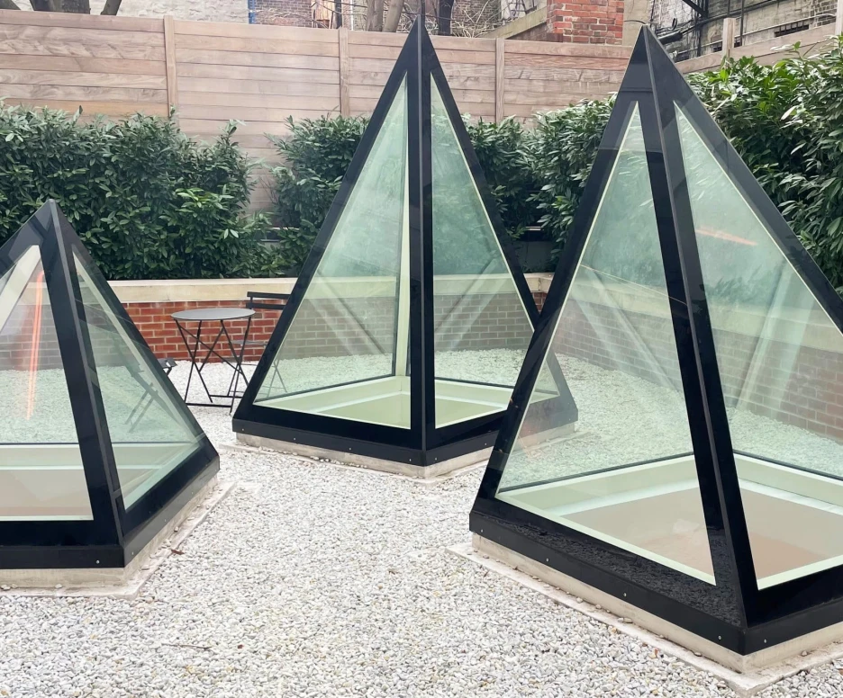 Three pyramid-shaped glass structures sit on a gravel surface, surrounded by low greenery and a brick wall. A small black table is visible nearby.