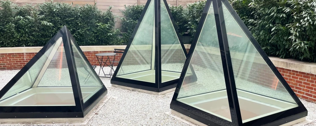Three glass pyramids with black frames are situated in a gravel area surrounded by greenery and a low brick wall. A small table and chair are visible in the background.