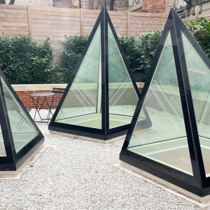Three pyramid-shaped glass structures sit on a gravel surface, surrounded by low greenery and a brick wall. A small black table is visible nearby.