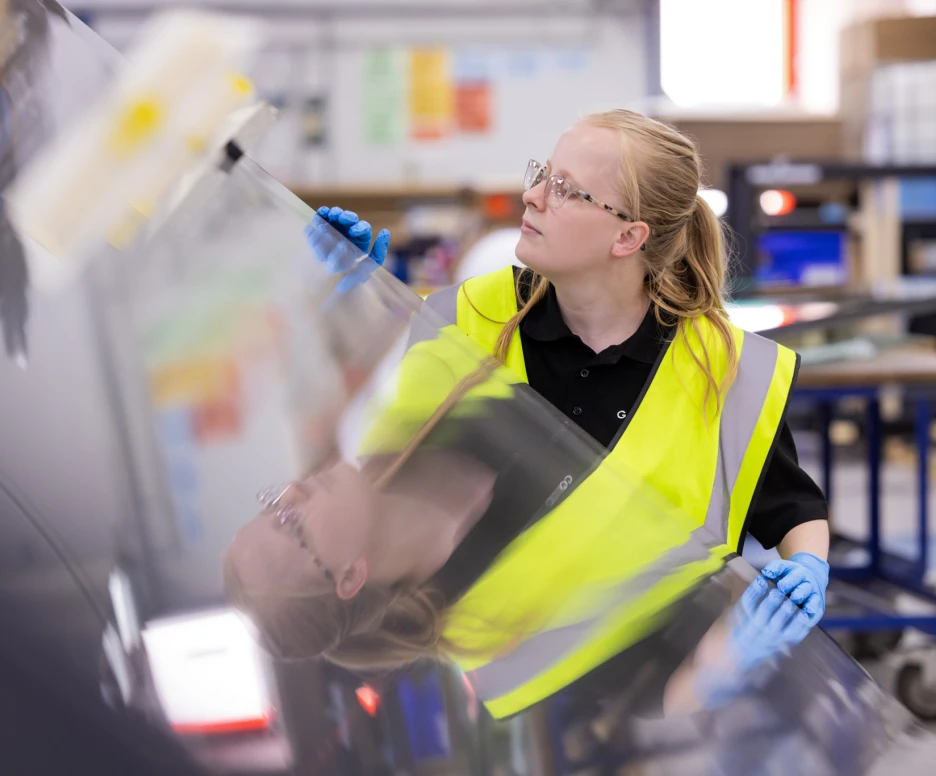 A person wearing a yellow safety vest and blue gloves inspects a shiny, reflective surface in a workshop. The individual has long blonde hair tied back and is focused on their task.