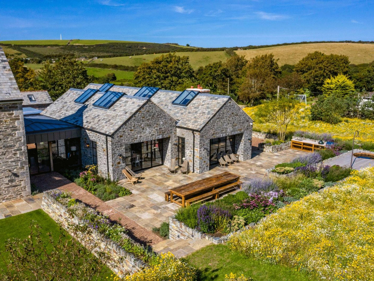 A modern stone house with a grey slate roof, surrounded by a vibrant garden filled with yellow wildflowers and greenery. A spacious patio features wooden dining tables and chairs, with hills in the background under a clear blue sky.